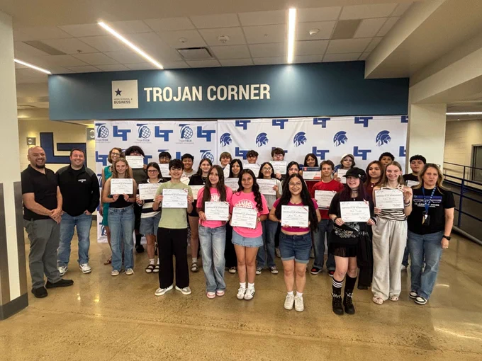 A large group of approximately 30 high school students and several adults gather in front of the Trojan Corner at Longmont High School; most students are holding Certificates of Achievement. Two adults stand to the left and one to the right.