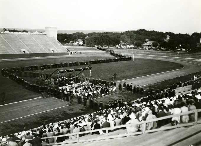 An outdoor graduation ceremony at a stadium with attendees seated in the stands and people in academic robes walking in a procession on the field.