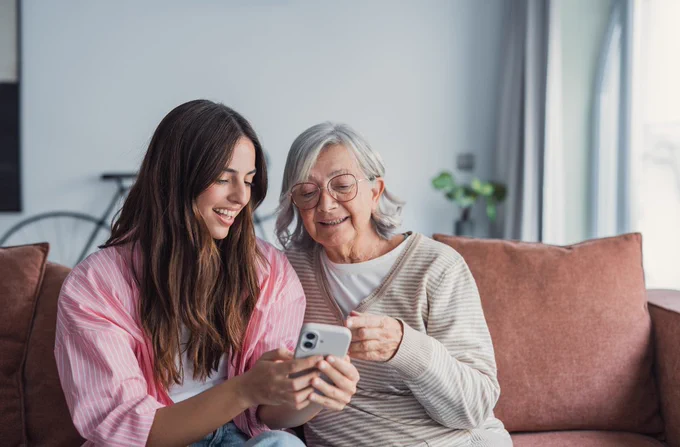 Two people seated on a couch, smiling while looking at a smartphone. The room has a cozy ambiance with a bicycle in the background and a vase with green plants on the table.