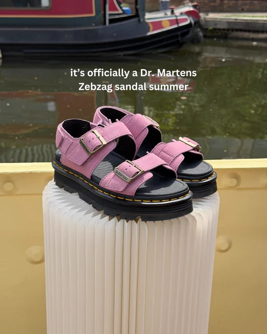 An image of a pair of pale pink Dr Martens Zebzag sandals sitting on a white podium next to a canal.