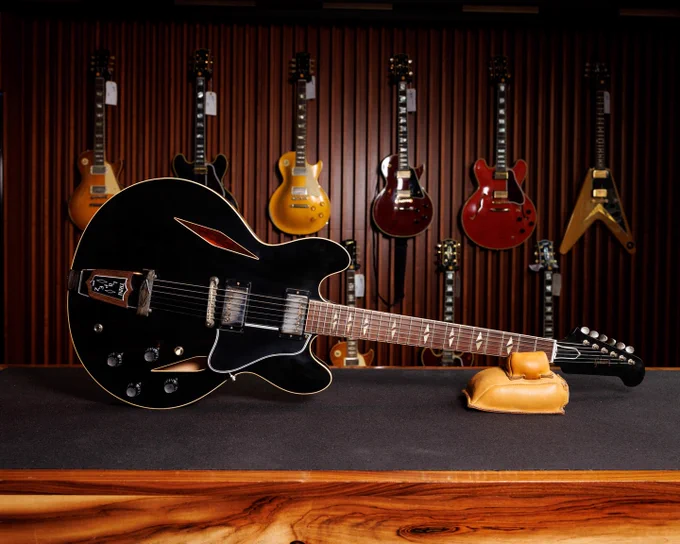 A black Gibson Trini Lopez electric guitar with f-holes is displayed on a stand in the foreground. Behind it, several colorful electric guitars hang on a wooden wall.