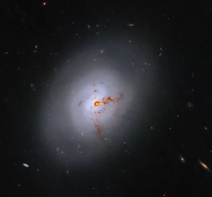 This Hubble image is of a bright lenticular galaxy (NGC 1266) seen nearly face on. Broad bright and dimmer areas of light hint at spiral structure, but there are no distinct spiral arms. Rusty-reddish-brown dust bisects the galaxy. More distant galaxies dot the black background, some even shining through the less dense regions of the bright foreground lenticular galaxy.