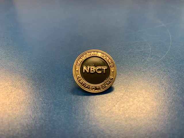Picture of a national board pin sitting on a table. The pin get pinned to the recipients shirt when they become certified as a National Board Certified Teacher.