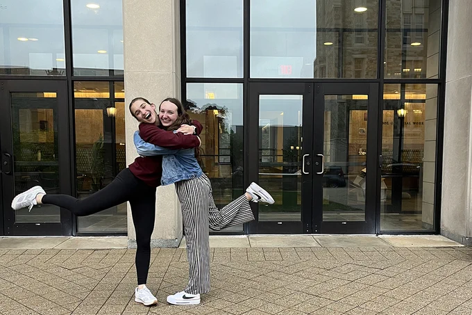 Two people joyfully embrace and pose with one leg raised in front of a building with large glass doors.