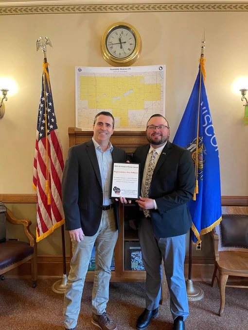 Representative Alex Dallman (right, in a dark suit and patterned tie) and Larry from the Wisconsin Towns Association (left, in a blazer and khaki pants) stand side-by-side smiling in an office. Representative Dallman holds the 2025-26 Friend of Towns Award plaque. They are positioned in front of a large map of Wisconsin Assembly District 39, with an American flag on the left and a Wisconsin state flag on the right.