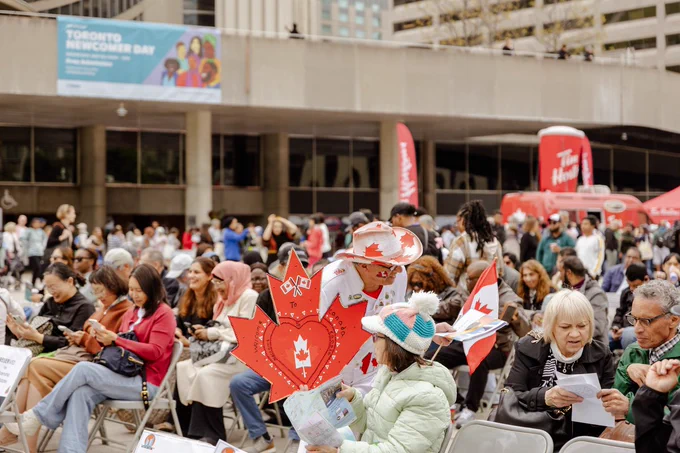 A large seated crowd gathers and is seated in front of the main stage at Nathan Phillips Square for Toronto Newcomer Day. In the foreground, attendees sit on folding chairs holding papers and small Canadian flags