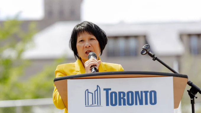Mayor Olivia Chow stands at an outdoor podium marked with the City of Toronto logo, speaking into a microphone. She wears a bright yellow jacket and is framed from the chest up. 