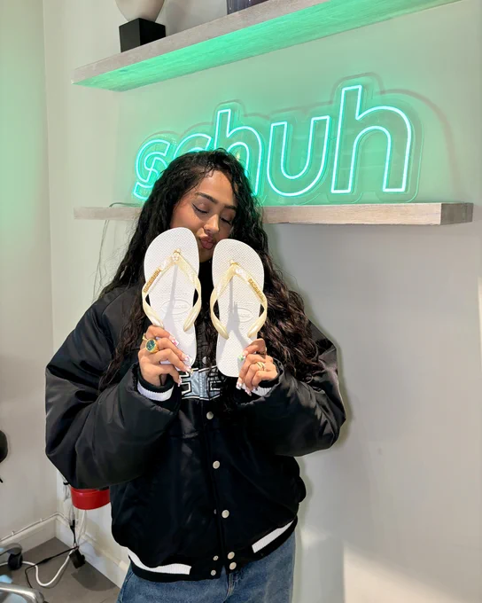 An image of a person holding a pair of white Havaianas Logo Metallic Flip flops whilst standing in front of a green neon sign that says "schuh".