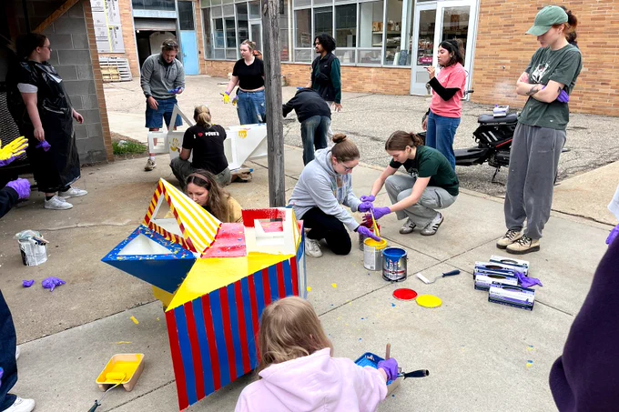 A group of people, some wearing gloves, paint colorful designs on large wooden structures outdoors. The scene is lively and collaborative.
