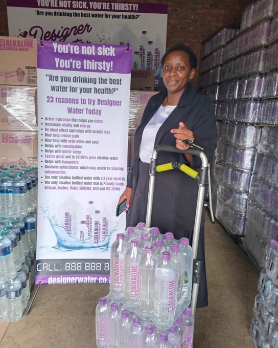 Cindy Hadio from the Alberton area recently joined as a Waterpreneur. She is seen smiling standing next to stacks of Designer Water pH10 in a warehouse.