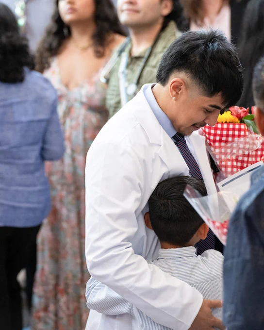 A person in a white coat is hugging a child while holding flowers. Other people are gathered in the background, with some looking on and one individual wearing a stethoscope.