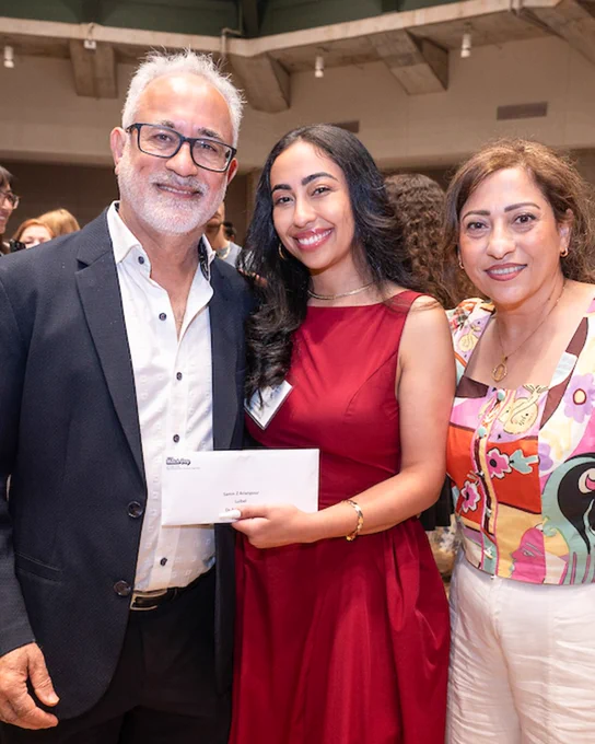 A group of three people smiling and posing indoors at an event. The person in the center holds an envelope and wears a red dress. The person on the left wears glasses and a dark suit, while the person on the right is dressed in a colorful top and light pants. Other attendees are visible in the background.
