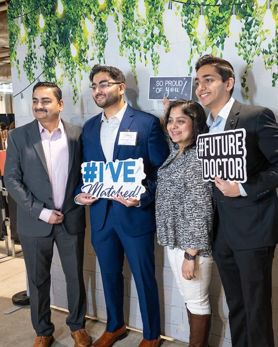 Four individuals are posing together in front of a decorative backdrop with hanging greenery and lights. Two people are holding signs that read "#I've Matched!" and "#Future Doctor." A small sign in the background says "So proud of you!" They are dressed in formal and business casual attire.
