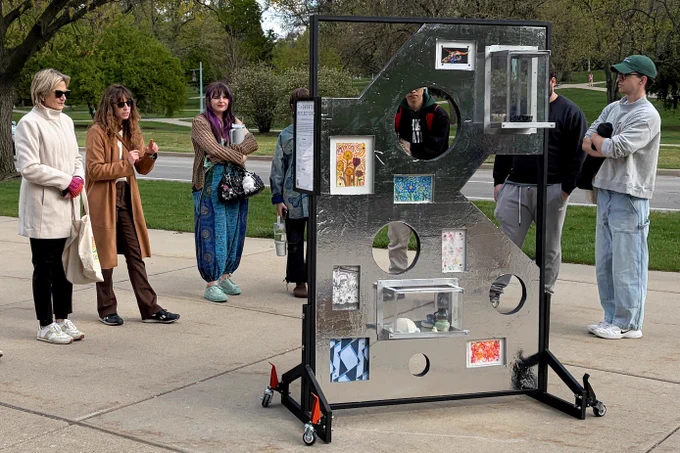 A group of people stands outdoors around a metal art display with various pictures and objects placed in circular cutouts. The mood is curious and engaging.