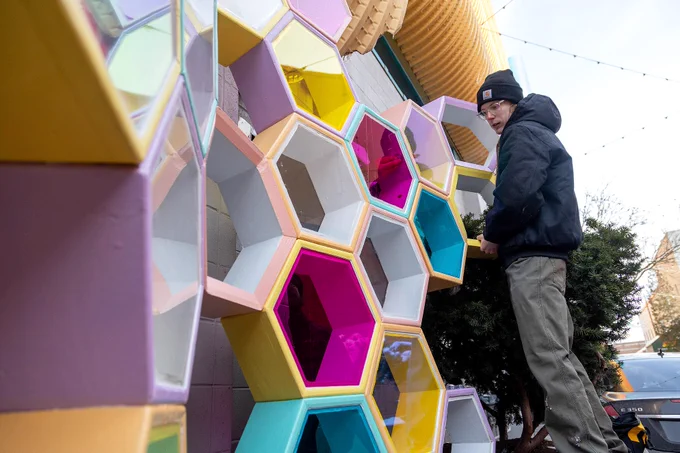 A person stands next to a vibrant, multicolored honeycomb art installation featuring hexagonal sections in pink, yellow, and teal. The mood is creative and lively.