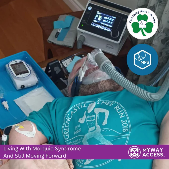 Dermot Devlin receiving his weekly Vimizim treatment for Morquio syndrome while lying on a sofa at home. He is wearing a green Greencastle 5 Mile Run t-shirt and using breathing support equipment connected to a ventilator machine beside him. Medical equipment and monitoring devices are visible nearby. The image includes MPS awareness logos and a My Way Access banner reading “Living With Morquio Syndrome And Still Moving Forward.”