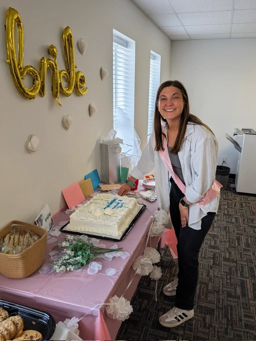 A woman is standing by a table, smiling and cutting a cake. The word "bride" is on a wall by the table.