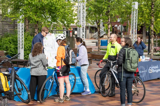 A group of people with bicycles are gathered around a map on an outdoor table. The table has a blue cover with "Montgomery Planning" printed on it. The scene takes place in a plaza with trees and brick paving.