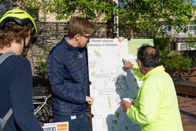 Two people are outside examining a map labeled "Master-planned bikeways in Wheaton." One person is pointing at the map, while another observes. A bicycle and trees are visible in the background.
