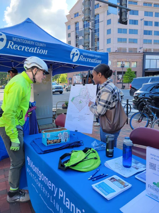 A person in bright cycling gear stands next to a Montgomery County Recreation tent, engaging with a person holding a map. Various items, including brochures and water bottles, are displayed on a table in front of them. Buildings and a street are visible in the background.