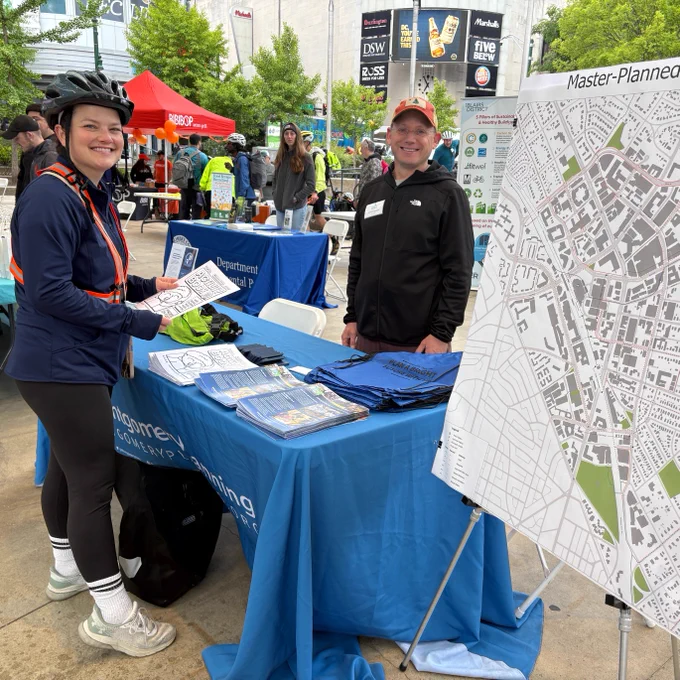 A person with a bike helmet is standing next to an information table at outdoor community event smiling at the camera next to a person manning the information table with a poster board of map next to them.