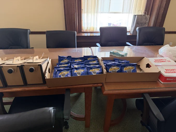 Cardboard canteens of coffee next to boxes filled with breakfast sandwiches and 2 boxes of doughnuts from Bogart's Donuts on a table with conference chairs around it. 
