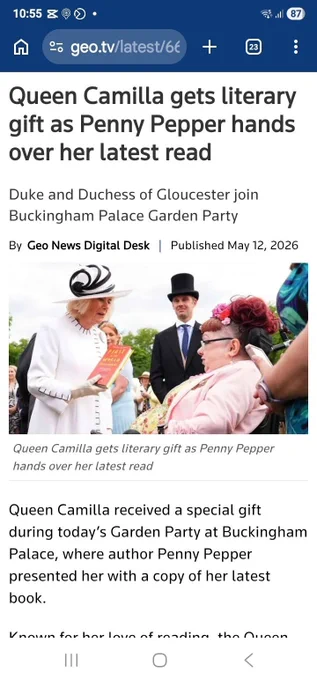 A screenshot of a photo showing Penny meeting Queen Camilla at an outdoor garden party at Buckingham Palace. Queen Camilla wears a black and white hat and a white coat with black detail. She’s holding Penny’s memoir, First in the World Somewhere. Penny sits in her wheelchair, facing the Queen. She is wearing a pale pink jacket, and has flowers in her red hair. The image is outdoors, bright, distant trees, and in the background stands a man in a top hat.