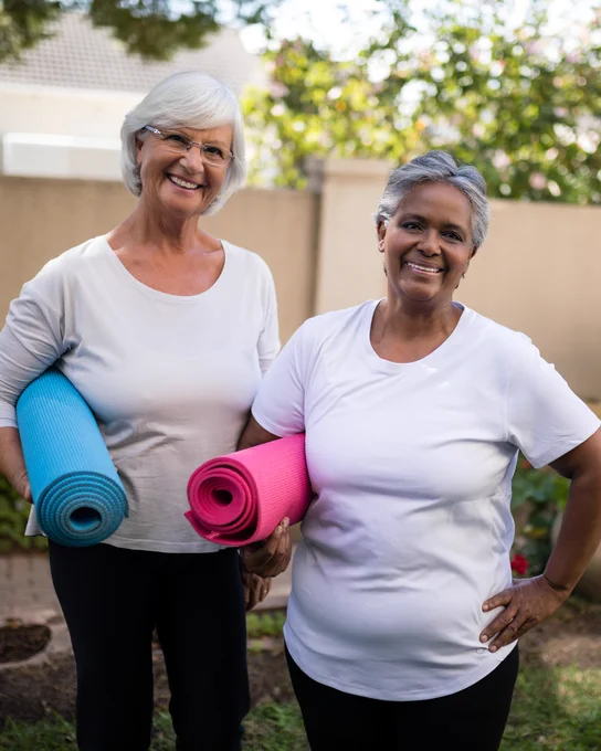 Photo Description: Two senior women standing outside with yoga mats in hand. 