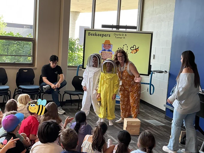 A group of children seated on the floor, watching presenters dressed as bees and flowers in front of a Beekeepers exhibit sign.