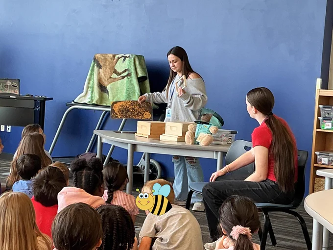 A woman is seated at a table in a classroom, surrounded by young students. Various objects, about beekeeping, are present on the table.