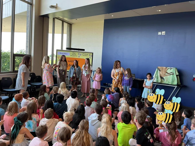 Large group of children and teachers on a stage in a classroom setting, with colorful decorations and a large screen in the background.