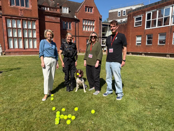 A posed shot of 4 adults facing camera and smiling on the Chief Constable's lawn at Police HQ in Guildford. From left to right as looking at image, there is Commissioner Lisa Townsend who is a blonde short haired lady.  She is wearing cropped cream tapered trousers and a light blue long sleeved top.  Next to her is PC Lucy Lucking who is wearing full police uniform. Then beside her is Kika the brown and white haired explosives detection dog who has a tennis ball in her mouth.  Then, from Kika, is Paula who is wearing a dark long sleeved green top and black trousers with a lanyard around her neck, and finally a tall man is beside her wearing light stonewash jeans and a black tee shirt with a lanyard around his neck.  In front of them on the lawn is around 15 tennis balls on the grass.