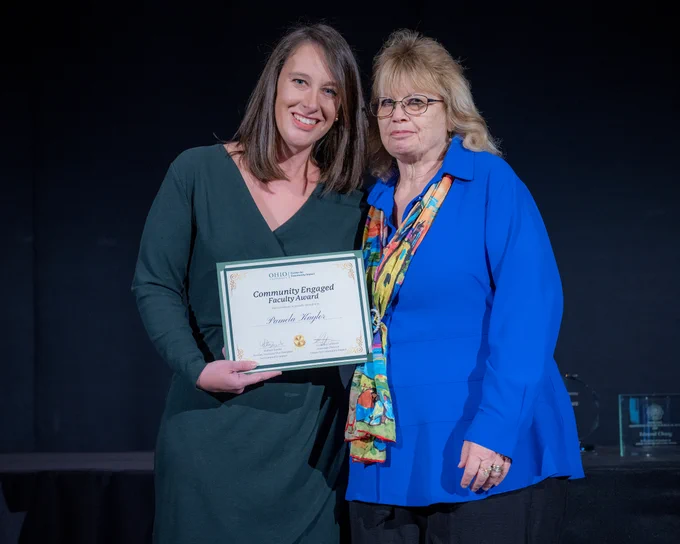 Pamela Kaylor poses with a certificate with another Ohio University faculty member in front of a black curtain.