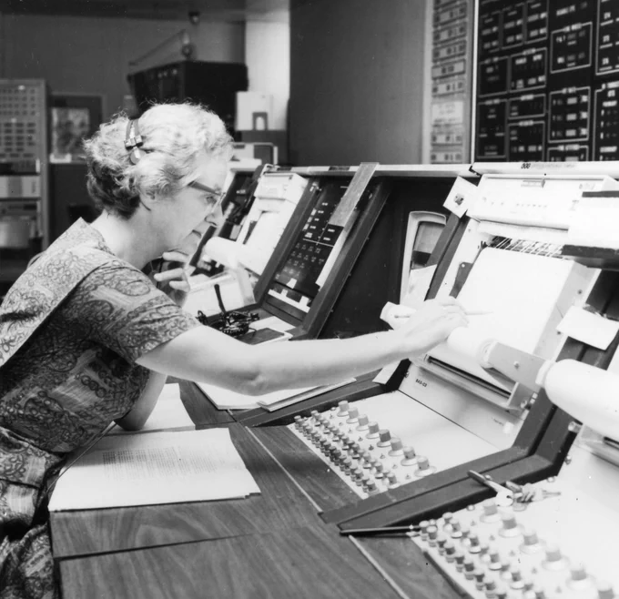 A black and white photo of Dr. Nancy Grace Roman sitting at a booth in a control console at NASA\u2019s Goddard Space Flight Center.  She has her right arm extended over the desk in front of her, pointing at some data on the console. The angled top of the desk is covered in instrumentation and buttons. 