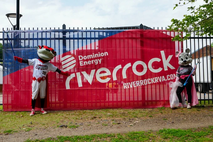 Two mascots are posing at the Dominion Energy Riverrock event, standing in front of a branded banner. The scene is lively and playful.