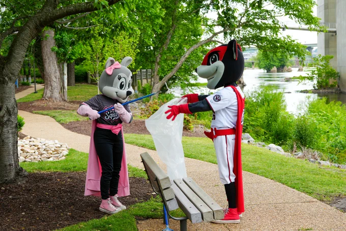 Two costumed mascots are gathering trash in a park, promoting environmental awareness and teamwork. The scene is bright and cheerful.