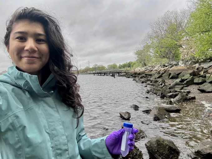 Shailja Gangrade holding a water sample near a rocky shoreline at India Point Park.