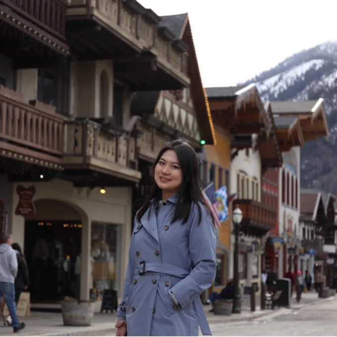 A person wearing a blue coat stands on a quaint, European-style street in Leavenworth, with snow-capped mountains in the background. The scene features charming wooden buildings and other people walking in the distance.