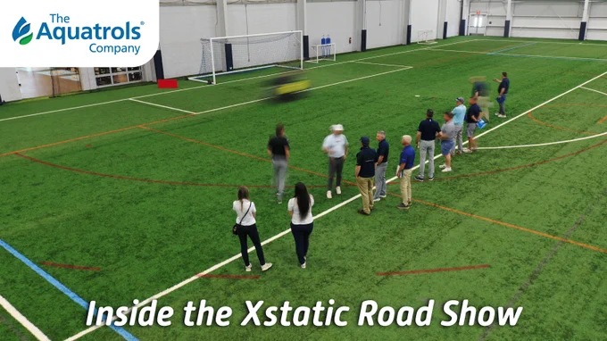 A group of people standing on an indoor soccer field near a goal. The Aquatrols Company logo is visible in the top left corner. Text reads: "Inside the Xstatic Road Show."