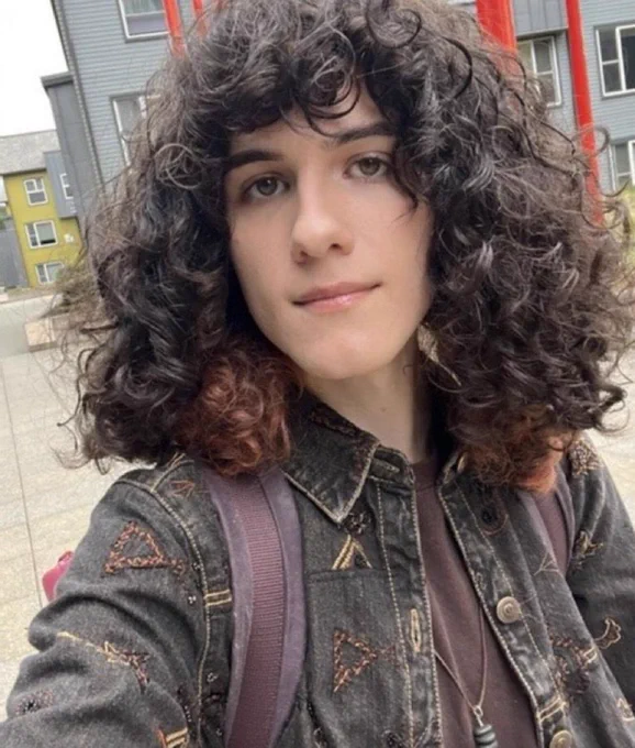 A selfie of Juniper Blessing. They have curly dark brown hair, pale skin, brown eyes, and are wearing a black denim jacket and brown shirt underneath with 2 dark purple backpack straps visible over their jacket.