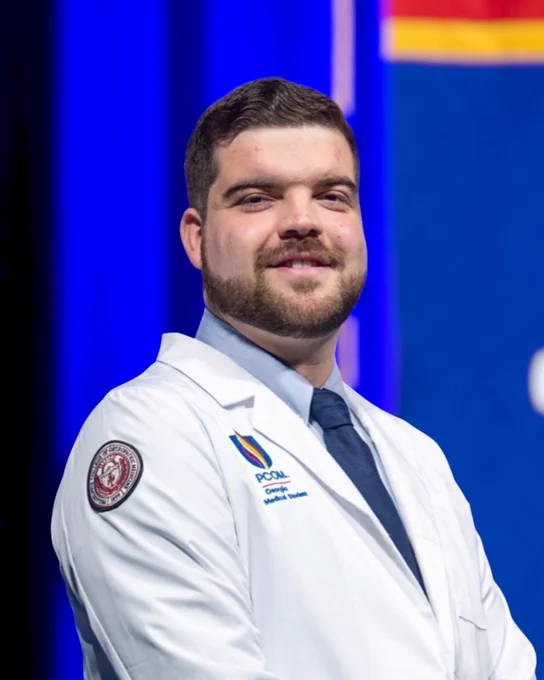 A person in a white coat with a badge and "PCOM" logo stands against a blue background.