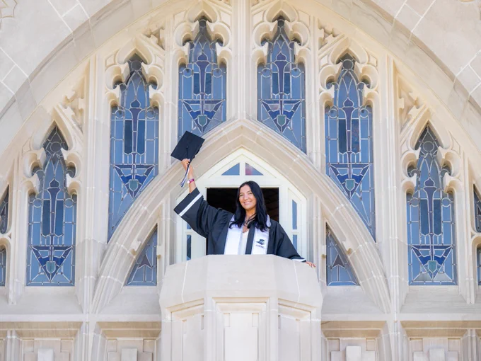 A person in graduation attire stands at a window, holding a graduation cap aloft. The background features Gothic-style architecture and stained glass windows.