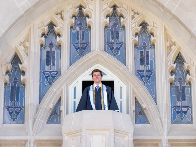 A person in graduation attire stands in an arched stone balcony beneath decorative stained glass windows.