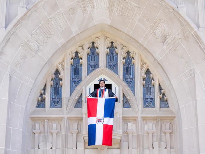 A person in graduation attire stands holding the Dominican Republic flag in front of the entrance of a stone building.