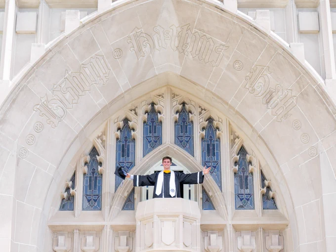 A person in graduation attire stands with arms spread wide on a Gothic-style building's balcony. The building features ornate arched windows in the background.