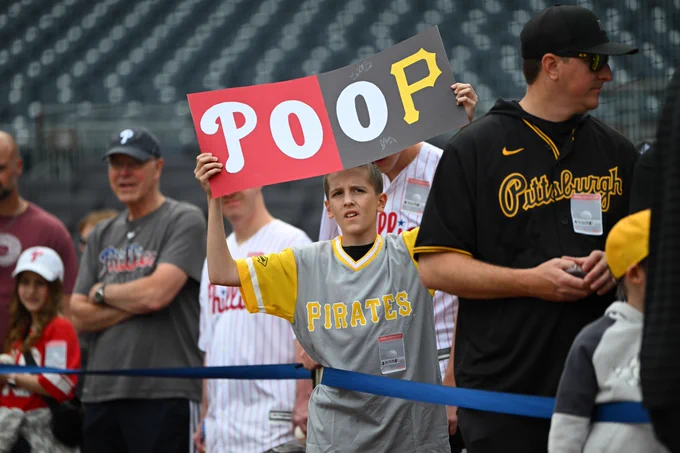 A Pirates fan holds a poster with an old score bug from a Phillies/Pirates brodcast.
It shows the Phillies logo with 0 runs scored, followed by 0 more runs scored and a Pirates logo.