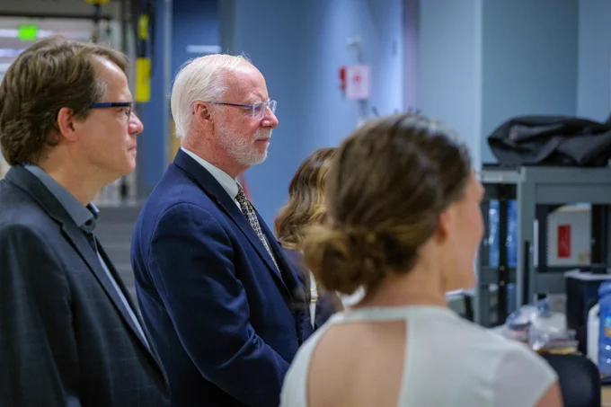 JB stands with a group listening attentively in a modern office setting.