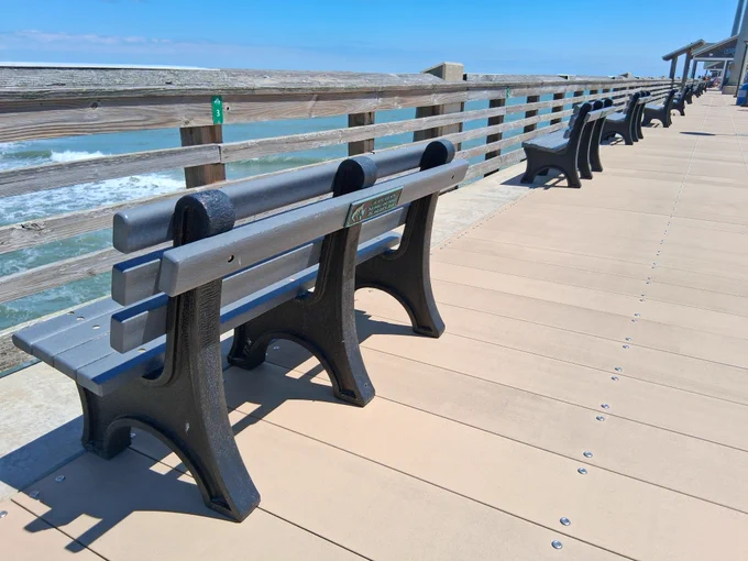 A boardwalk lined with empty benches overlooks the ocean. Waves are visible in the background, and a clear blue sky stretches above.