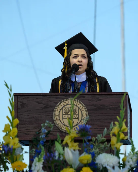 Gulsinam Akhmadzhanova, wearing a graduation cap and gown and speaking from the podium.