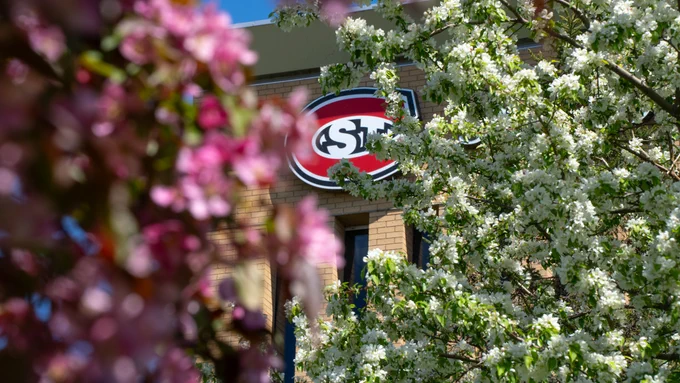 Blooming trees with pink and white flowers partially obscure a building with an SCSU sign in the background.